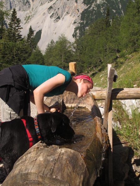 Kristina trinkt zusammen mit Hund an einem Brunnen in den Bergen
